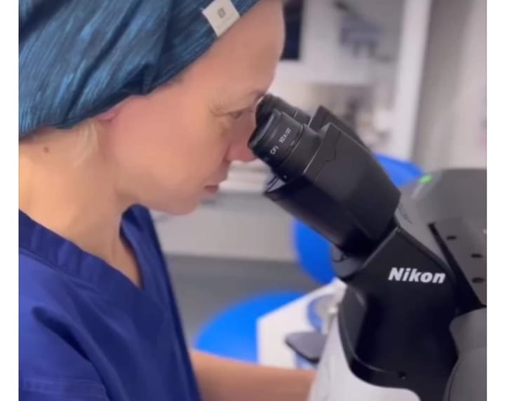 A woman in scrubs looking down a microscope.